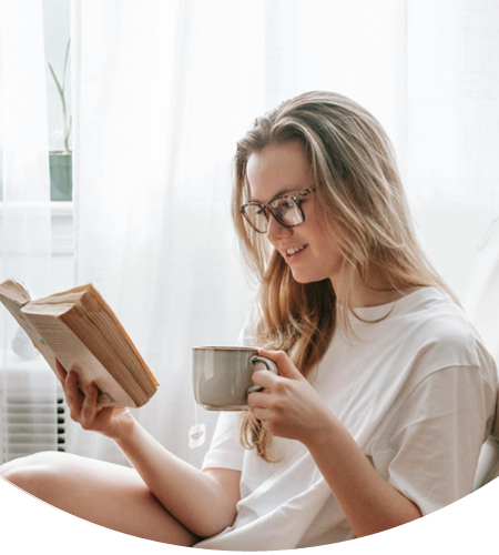 Junge Frau sitzt mit Lesebrille im Sessel, hält eine Tasse und liest ein Buch